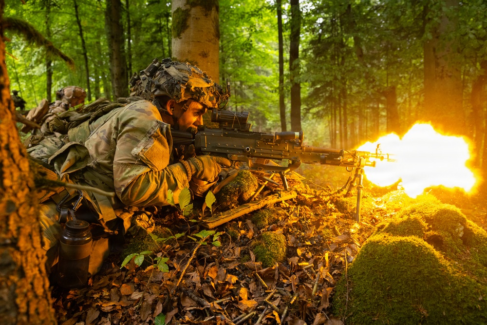 A soldier in full camouflage gear is prone in a forest setting, firing a light machine gun with a visible muzzle flash. The weapon appears to be mounted on a bipod and is actively discharging, producing a large orange flame at the muzzle. At least one additional soldier is visible in the background, suggesting a squad-level exercise or operation.