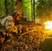 A soldier in full camouflage gear is prone in a forest setting, firing a light machine gun with a visible muzzle flash. The weapon appears to be mounted on a bipod and is actively discharging, producing a large orange flame at the muzzle. At least one additional soldier is visible in the background, suggesting a squad-level exercise or operation.