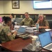 A formal military conference room meeting with a mix of uniformed military personnel and civilian attendees seated around a large wooden conference table. Multiple participants have laptops open and name placards are visible in front of several seats. Two wall-mounted monitors and a dual-timezone digital clock display are visible in the background.