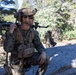 A U.S. Marine in full combat gear is kneeling on a large rock in a forested, mountainous environment, holding a rifle across his shoulder while scanning his surroundings. He is equipped with a tactical helmet, plate carrier vest, and multiple pouches. Additional personnel in similar uniforms are visible in the background.