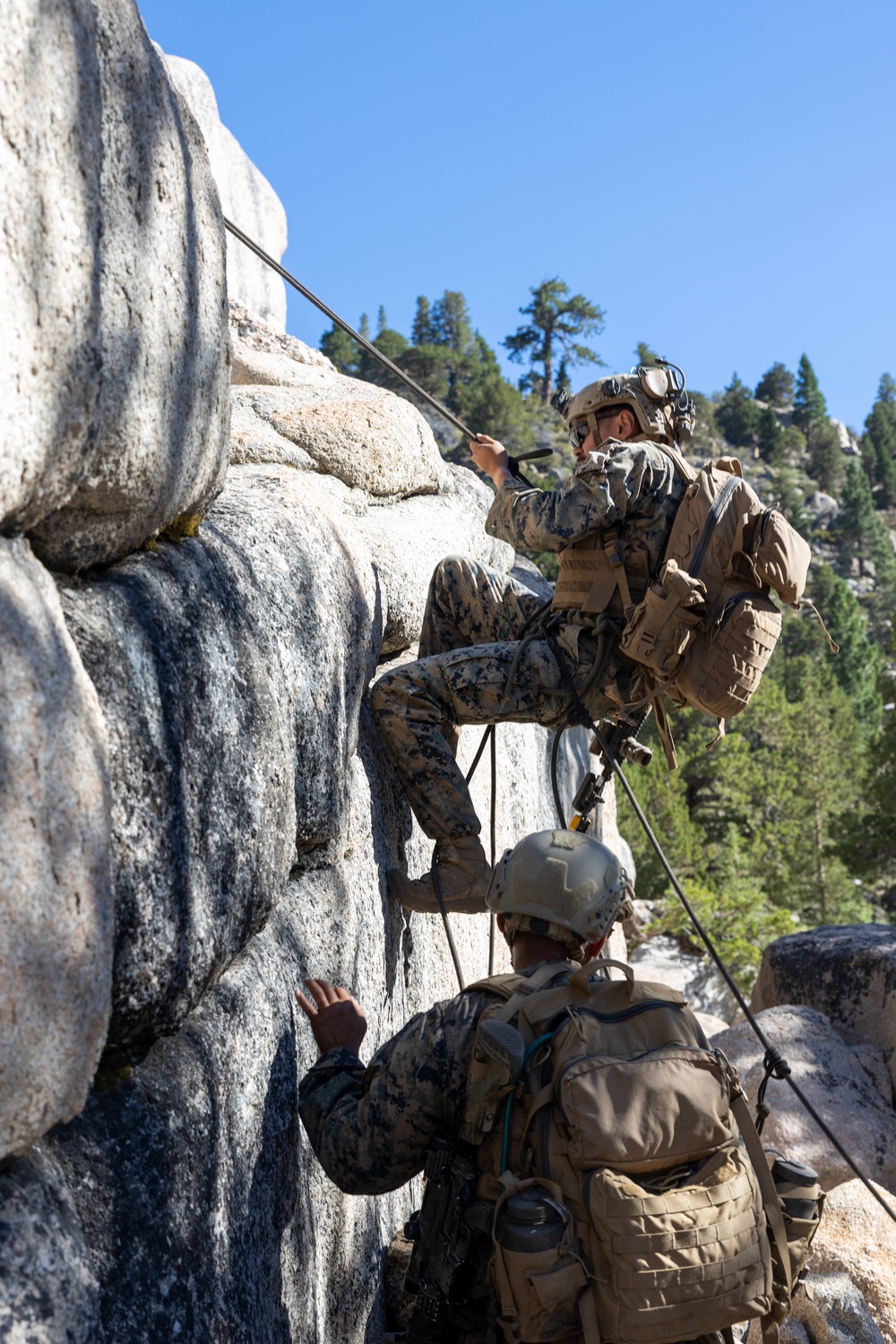 Two military personnel in camouflage uniforms are ascending a granite rock face in a mountainous, forested environment. The upper climber is equipped with full tactical gear including a helmet, backpack, and what appears to be a radio antenna, while using ropes for the ascent. This appears to be a military mountain warfare or rock climbing training exercise.