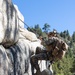 Two military personnel in camouflage uniforms are ascending a granite rock face in a mountainous, forested environment. The upper climber is equipped with full tactical gear including a helmet, backpack, and what appears to be a radio antenna, while using ropes for the ascent. This appears to be a military mountain warfare or rock climbing training exercise.