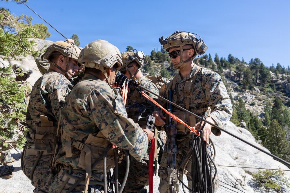 A group of military personnel in MARPAT camouflage uniforms are gathered on a rocky outcrop in a mountainous, forested environment. They appear to be conducting rope or rigging work, handling multiple lines including orange and black ropes. The setting and equipment suggest a mountain warfare or rappelling training exercise.