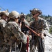 A group of military personnel in MARPAT camouflage uniforms are gathered on a rocky outcrop in a mountainous, forested environment. They appear to be conducting rope or rigging work, handling multiple lines including orange and black ropes. The setting and equipment suggest a mountain warfare or rappelling training exercise.