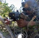 A military or law enforcement operator is positioned in dense brush, aiming a rifle equipped with tactical accessories including an optic scope and what appears to be a laser/illuminator device. The individual is wearing a ballistic helmet with a mounted device (likely a night vision mount), tactical sunglasses, and camouflage uniform. The scene suggests a field training exercise or operational reconnaissance in a natural outdoor environment.