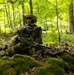 Two soldiers in full combat gear are positioned in a deciduous forest, taking up a defensive or observation posture behind moss-covered rocks. The foreground soldier is prone with a light machine gun mounted on a bipod, while the second soldier kneels behind in an alert, scanning posture. The setting and equipment suggest a military training exercise in a temperate European forest environment.