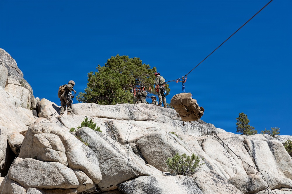 A group of military personnel in camouflage uniforms are conducting rope and rappelling training on a granite rock face under a clear blue sky. Multiple individuals are visible on the ridge, managing ropes and rigging systems, while a large tactical backpack is suspended mid-air on a rope line. The setting appears to be a rocky, arid mountainous environment with sparse coniferous trees.