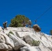 A group of military personnel in camouflage uniforms are conducting rope and rappelling training on a granite rock face under a clear blue sky. Multiple individuals are visible on the ridge, managing ropes and rigging systems, while a large tactical backpack is suspended mid-air on a rope line. The setting appears to be a rocky, arid mountainous environment with sparse coniferous trees.