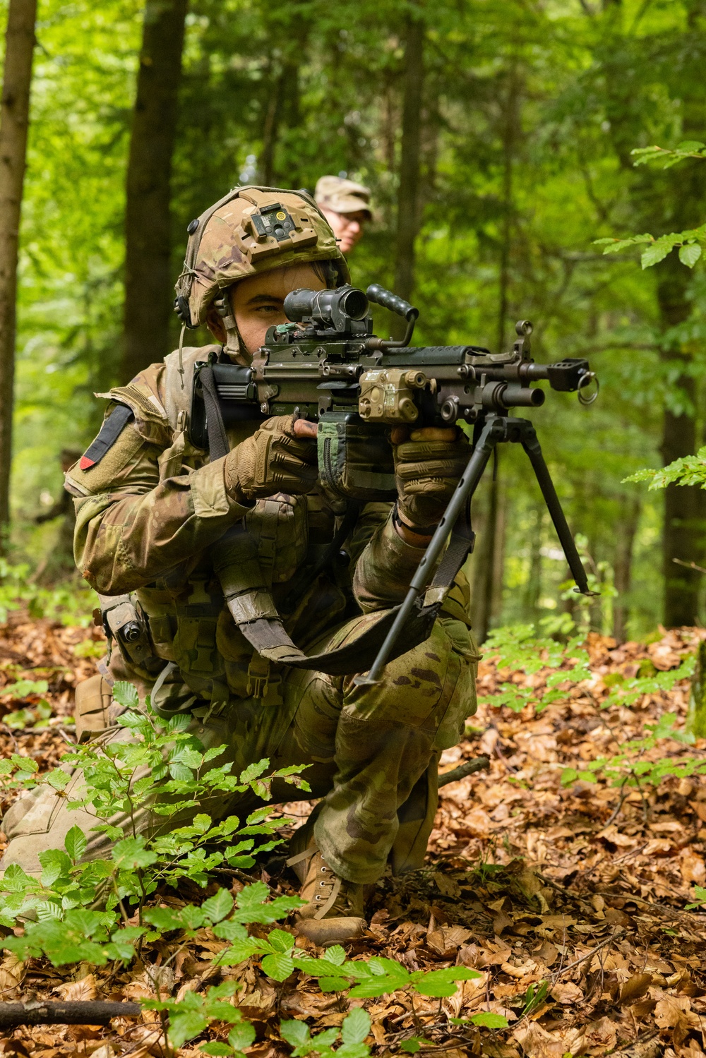 A soldier in full combat gear is crouched in a forested environment, aiming a light machine gun equipped with a bipod and optical scope. A second individual wearing a patrol cap is visible in the background. The setting and equipment suggest a military training exercise in a deciduous forest.