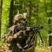 A soldier in full combat gear is crouched in a forested environment, aiming a light machine gun equipped with a bipod and optical scope. A second individual wearing a patrol cap is visible in the background. The setting and equipment suggest a military training exercise in a deciduous forest.
