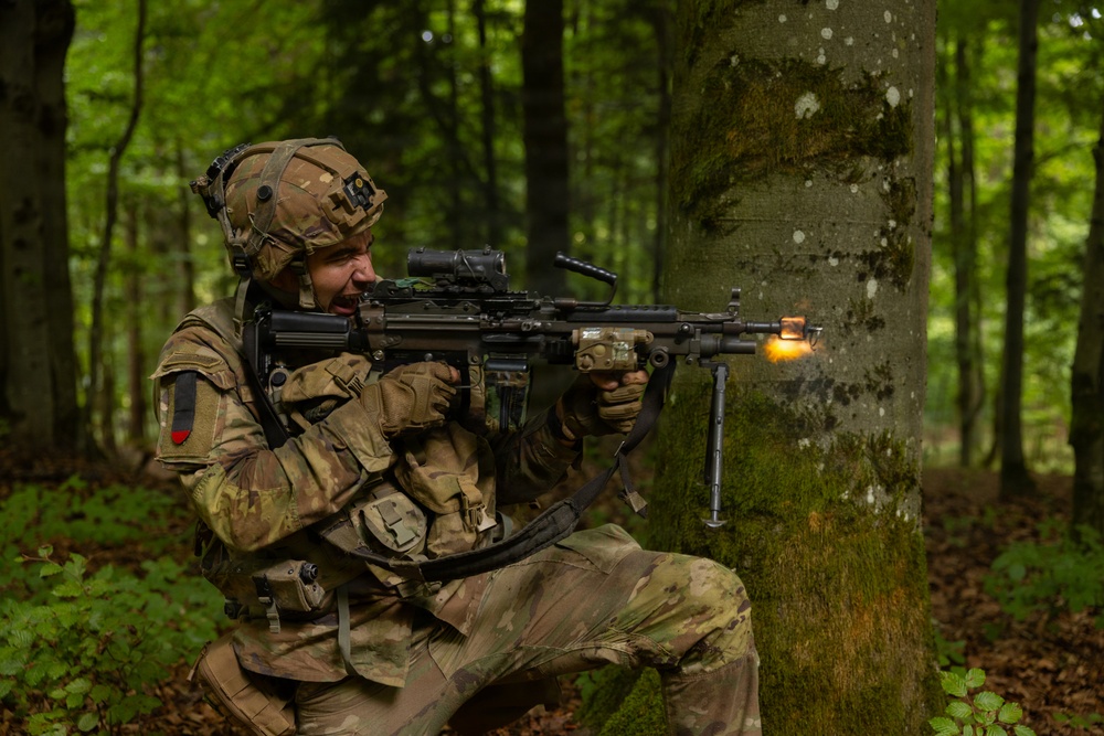 A soldier in full multicam camouflage gear is firing a light machine gun from a kneeling position against a tree in a deciduous forest. A muzzle flash is visible at the barrel, indicating the weapon is being discharged. The soldier appears to be in a training exercise based on the controlled environment and professional photography.