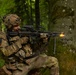 A soldier in full multicam camouflage gear is firing a light machine gun from a kneeling position against a tree in a deciduous forest. A muzzle flash is visible at the barrel, indicating the weapon is being discharged. The soldier appears to be in a training exercise based on the controlled environment and professional photography.