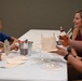 A family of four is seated around a white table engaged in a craft activity, assembling what appear to be small wooden structures. A metal bucket labeled 'USO' sits in the center of the table alongside craft supplies including glue, markers, and wooden pieces. The setting appears to be an indoor event space or community room.