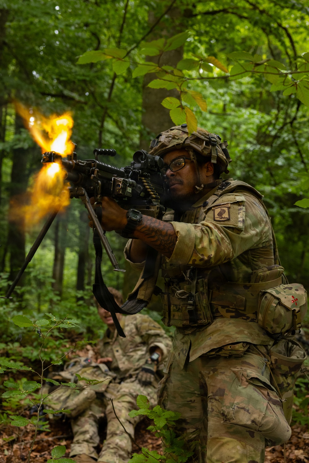 A U.S. Army soldier in Operational Camouflage Pattern (OCP) uniform fires a belt-fed machine gun (appears to be an M249 SAW or M240B) in a wooded environment, with a visible muzzle flash. Additional soldiers in matching uniforms are visible in the background in prone positions. This appears to be a military training exercise or live-fire drill in a forested setting.
