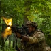 A U.S. Army soldier in Operational Camouflage Pattern (OCP) uniform fires a belt-fed machine gun (appears to be an M249 SAW or M240B) in a wooded environment, with a visible muzzle flash. Additional soldiers in matching uniforms are visible in the background in prone positions. This appears to be a military training exercise or live-fire drill in a forested setting.