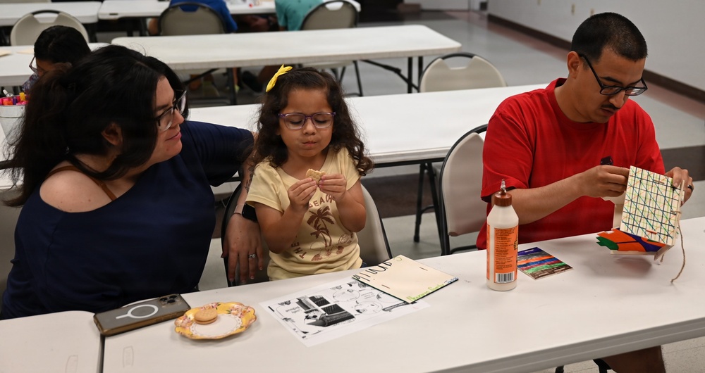 A family of three — a woman, a young girl, and a man — are seated at a white folding table in what appears to be a community center or church hall. The young girl is examining a small food item while the man holds up a colorful handmade craft project. The setting suggests a community arts and crafts event or workshop.