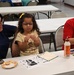A family of three — a woman, a young girl, and a man — are seated at a white folding table in what appears to be a community center or church hall. The young girl is examining a small food item while the man holds up a colorful handmade craft project. The setting suggests a community arts and crafts event or workshop.