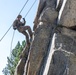 A military service member in full camouflage uniform and helmet is rappelling down a steep rocky cliff face using ropes. The individual appears to be engaged in a military training exercise involving vertical rope descent techniques. The setting is an outdoor rocky terrain with coniferous trees and a clear blue sky visible in the background.