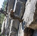 Two military personnel in camouflage uniforms and helmets are rappelling down a steep granite rock face in a mountainous, forested environment. The scene appears to be a military training exercise involving vertical rope descent techniques. Tall conifer trees and a partially visible mountain peak are visible in the background.
