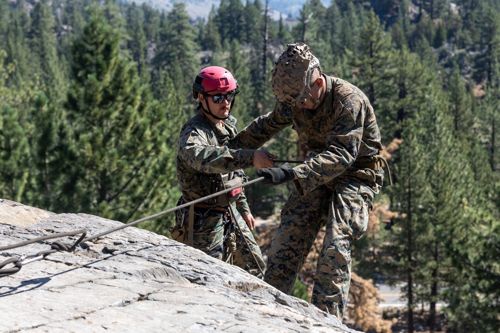 Two military personnel in camouflage uniforms are engaged in rappelling or rope training on a granite rock face. One individual wearing a red climbing helmet and sunglasses appears to be instructing or assisting the other, who is wearing a camouflage patrol cap. The setting is a mountainous, forested environment with tall conifer trees visible in the background.