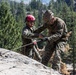 Two military personnel in camouflage uniforms are engaged in rappelling or rope training on a granite rock face. One individual wearing a red climbing helmet and sunglasses appears to be instructing or assisting the other, who is wearing a camouflage patrol cap. The setting is a mountainous, forested environment with tall conifer trees visible in the background.