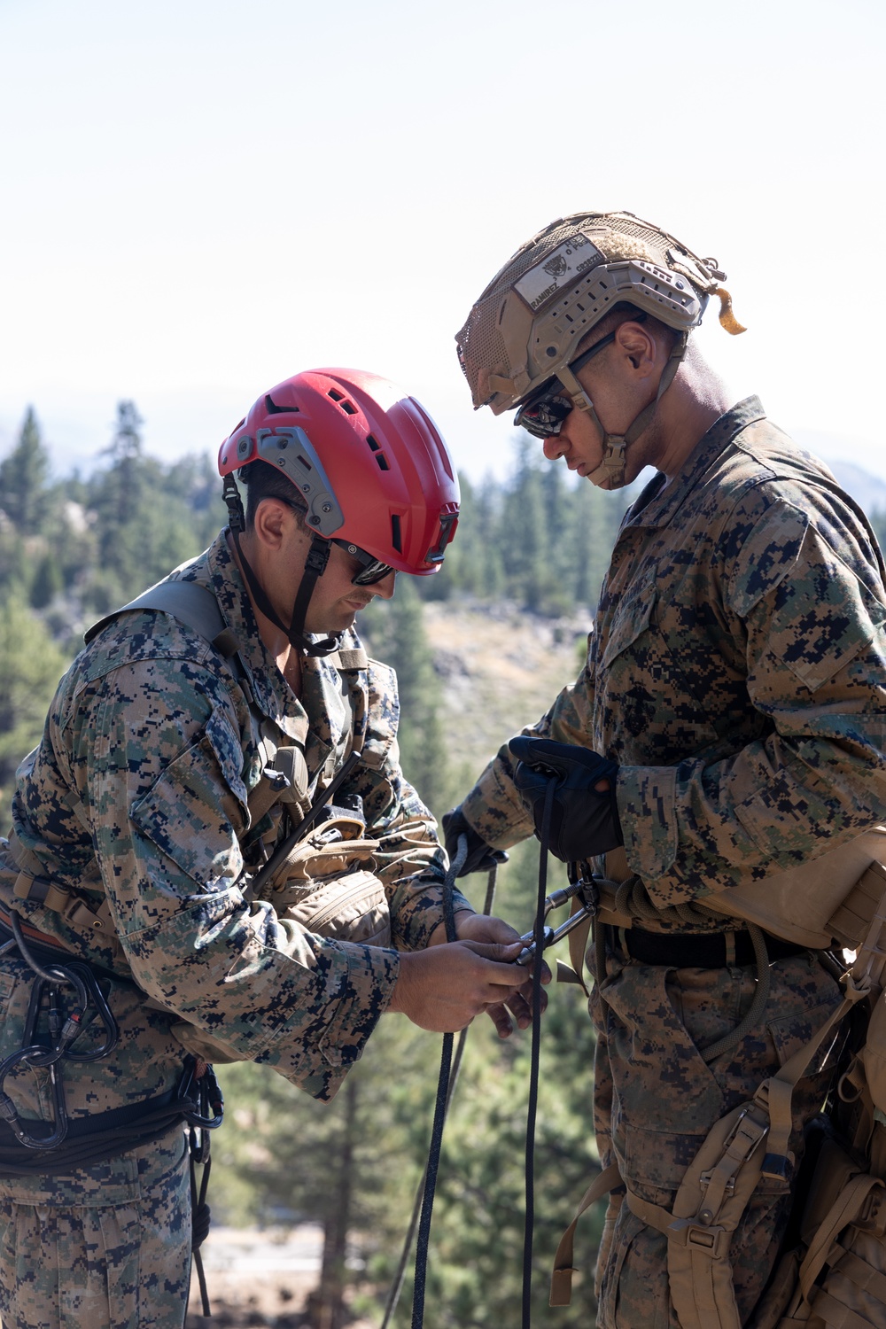 Two military personnel in digital camouflage uniforms are working together with climbing/rappelling rope and hardware in an outdoor mountainous environment. One individual wears a red civilian climbing helmet while the other wears a tan tactical military helmet with netting. They appear to be checking or rigging rope equipment, likely during a mountain warfare or rappelling training exercise.