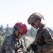 Two military personnel in digital camouflage uniforms are working together with climbing/rappelling rope and hardware in an outdoor mountainous environment. One individual wears a red civilian climbing helmet while the other wears a tan tactical military helmet with netting. They appear to be checking or rigging rope equipment, likely during a mountain warfare or rappelling training exercise.