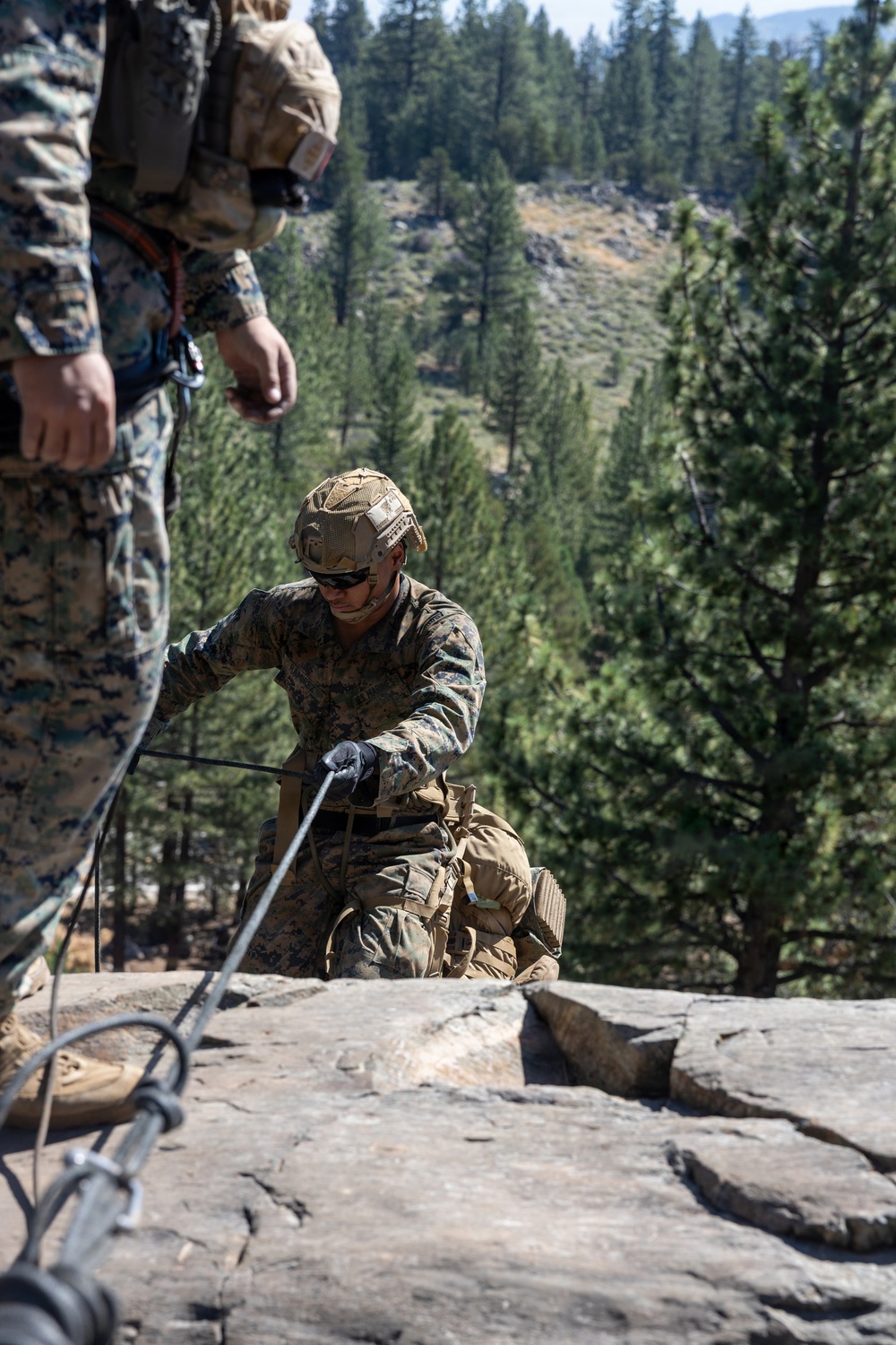 Two military personnel in MARPAT (Marine pattern) camouflage uniforms are conducting rock climbing or rappelling training on a granite rock face. The central figure is equipped with a tactical helmet, sunglasses, gloves, and full combat gear while managing ropes at the edge of a cliff. The background shows a forested mountain terrain with tall pine trees under clear daylight conditions.