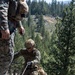 Two military personnel in MARPAT (Marine pattern) camouflage uniforms are conducting rock climbing or rappelling training on a granite rock face. The central figure is equipped with a tactical helmet, sunglasses, gloves, and full combat gear while managing ropes at the edge of a cliff. The background shows a forested mountain terrain with tall pine trees under clear daylight conditions.