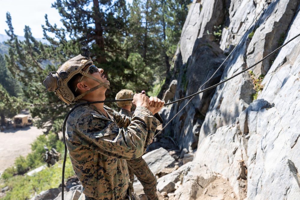 A military service member in digital camouflage uniform and tactical helmet is managing a rope at the base of a rocky cliff face, looking upward. At least one other service member is visible in the background. The setting appears to be a mountainous training environment with conifer trees and a valley visible below.