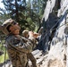 A military service member in digital camouflage uniform and tactical helmet is managing a rope at the base of a rocky cliff face, looking upward. At least one other service member is visible in the background. The setting appears to be a mountainous training environment with conifer trees and a valley visible below.