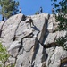 A group of military personnel in camouflage uniforms are conducting a rock climbing and rappelling training exercise on a large granite cliff face. Multiple individuals are positioned at various heights on the rock wall, with some at the summit acting as anchors or instructors, while others ascend or descend using ropes. The setting appears to be a mountainous, forested environment under clear blue skies.