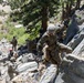 Military personnel in camouflage uniforms are conducting a rope-assisted rock climbing or rappelling training exercise on a rocky cliff face. Multiple soldiers are visible at various stages of the climb, with some on the rocks and others observing or managing ropes at the base. The setting appears to be a mountainous, forested training area during daylight hours.