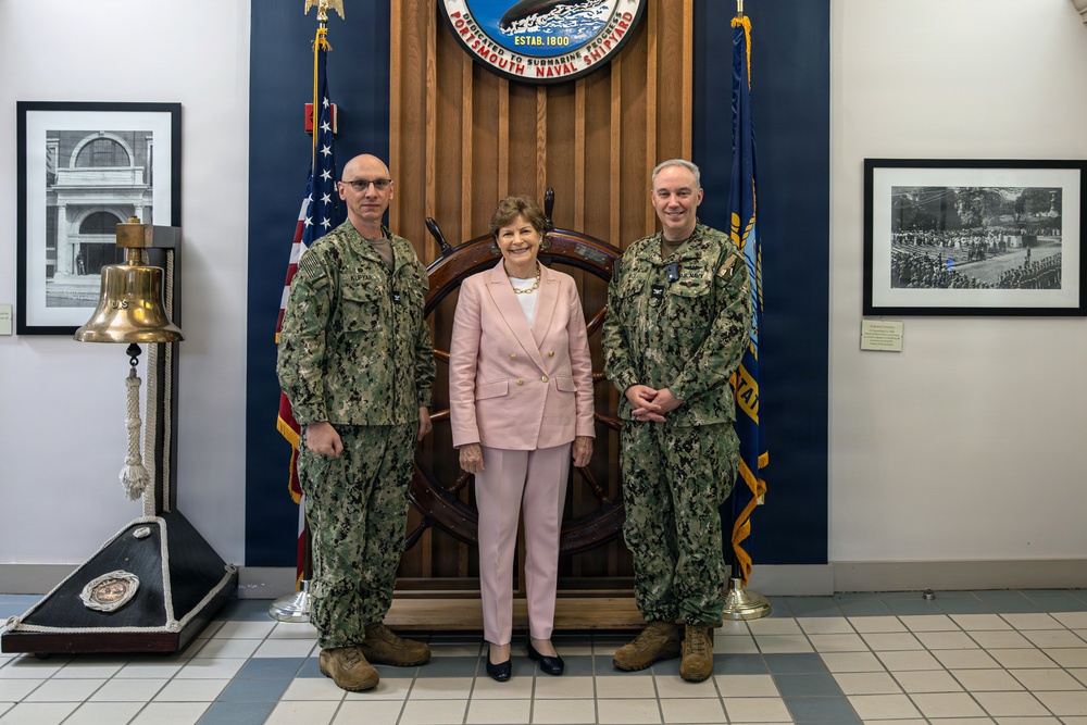 Three individuals pose for a formal photograph at Portsmouth Naval Shipyard, established in 1800, as indicated by the official seal visible on the wooden backdrop. Two personnel in U.S. Navy camouflage uniforms (NWU Type III) flank a civilian woman dressed in a pink suit. The setting appears to be an official reception or ceremonial area featuring nautical artifacts and historical photographs.