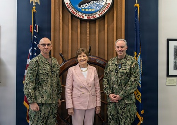 Three individuals pose for a formal photograph at Portsmouth Naval Shipyard, established in 1800, as indicated by the official seal visible on the wooden backdrop. Two personnel in U.S. Navy camouflage uniforms (NWU Type III) flank a civilian woman dressed in a pink suit. The setting appears to be an official reception or ceremonial area featuring nautical artifacts and historical photographs.