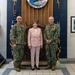 Three individuals pose for a formal photograph at Portsmouth Naval Shipyard, established in 1800, as indicated by the official seal visible on the wooden backdrop. Two personnel in U.S. Navy camouflage uniforms (NWU Type III) flank a civilian woman dressed in a pink suit. The setting appears to be an official reception or ceremonial area featuring nautical artifacts and historical photographs.