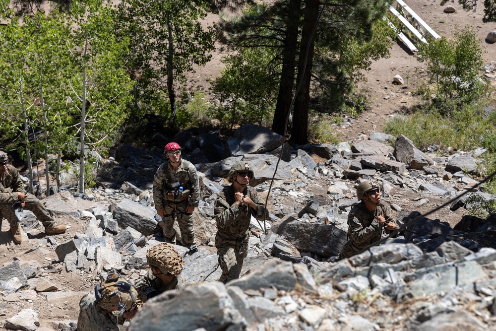 A group of military personnel in camouflage uniforms are conducting a rappelling or rope training exercise on a rocky mountainside. One individual wearing a distinctive red helmet appears to serve as an instructor or safety officer overseeing the activity. The terrain consists of loose shale and boulders with coniferous trees visible in the background.