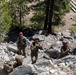 A group of military personnel in camouflage uniforms are conducting a rappelling or rope training exercise on a rocky mountainside. One individual wearing a distinctive red helmet appears to serve as an instructor or safety officer overseeing the activity. The terrain consists of loose shale and boulders with coniferous trees visible in the background.
