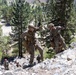 Military personnel in camouflage uniforms are conducting rappelling or climbing training on a rocky cliff face in a mountainous, forested environment. Two soldiers in the foreground are actively descending or traversing the rock face using ropes and harnesses, while additional personnel are visible at the base of the cliff. The setting appears to be a structured military mountain warfare or combat training exercise.