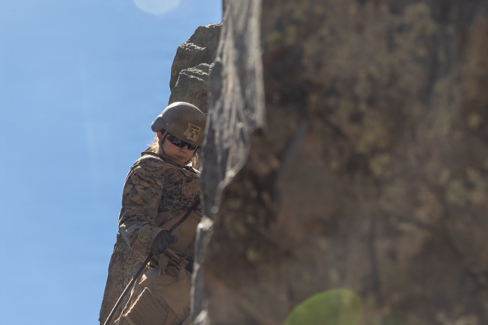A military service member in full combat gear is rappelling or climbing a steep rock face. The individual is wearing MARPAT (Marine pattern) camouflage uniform, a tactical helmet, sunglasses, and gloves, suggesting this is a military training exercise. The photo is taken from a low angle against a clear blue sky, emphasizing the height and scale of the rock formation.