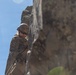 A military service member in full combat gear is rappelling or climbing a steep rock face. The individual is wearing MARPAT (Marine pattern) camouflage uniform, a tactical helmet, sunglasses, and gloves, suggesting this is a military training exercise. The photo is taken from a low angle against a clear blue sky, emphasizing the height and scale of the rock formation.