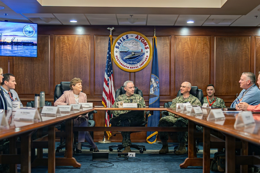 A formal meeting is taking place in a conference room at Portsmouth Naval Shipyard, as indicated by the large official seal on the wall reading 'Sails to Atoms - Portsmouth Naval Shipyard.' The gathering includes uniformed U.S. Navy personnel and civilian attendees seated around a large conference table. An American flag and a Navy command flag flank the official seal in the background.