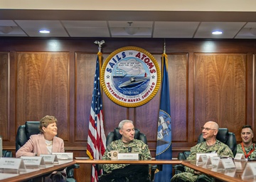 A formal meeting is taking place in a conference room at Portsmouth Naval Shipyard, as indicated by the large official seal on the wall reading 'Sails to Atoms - Portsmouth Naval Shipyard.' The gathering includes uniformed U.S. Navy personnel and civilian attendees seated around a large conference table. An American flag and a Navy command flag flank the official seal in the background.