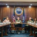 A formal meeting is taking place in a conference room at Portsmouth Naval Shipyard, as indicated by the large official seal on the wall reading 'Sails to Atoms - Portsmouth Naval Shipyard.' The gathering includes uniformed U.S. Navy personnel and civilian attendees seated around a large conference table. An American flag and a Navy command flag flank the official seal in the background.
