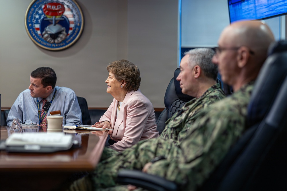 A formal meeting is taking place in what appears to be a naval command conference room, as indicated by the official seal visible on the wall. Four individuals are seated at a wooden conference table, including a civilian woman in a pink blazer and two military personnel in Navy camouflage uniforms. The setting suggests an official briefing or oversight visit at a naval installation.
