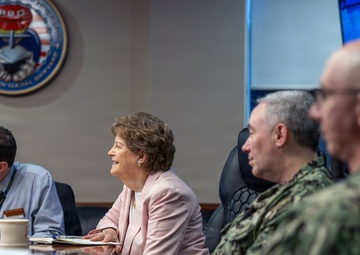 A formal meeting is taking place in what appears to be a naval command conference room, as indicated by the official seal visible on the wall. Four individuals are seated at a wooden conference table, including a civilian woman in a pink blazer and two military personnel in Navy camouflage uniforms. The setting suggests an official briefing or oversight visit at a naval installation.
