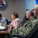 A formal meeting is taking place in what appears to be a naval command conference room, as indicated by the official seal visible on the wall. Four individuals are seated at a wooden conference table, including a civilian woman in a pink blazer and two military personnel in Navy camouflage uniforms. The setting suggests an official briefing or oversight visit at a naval installation.