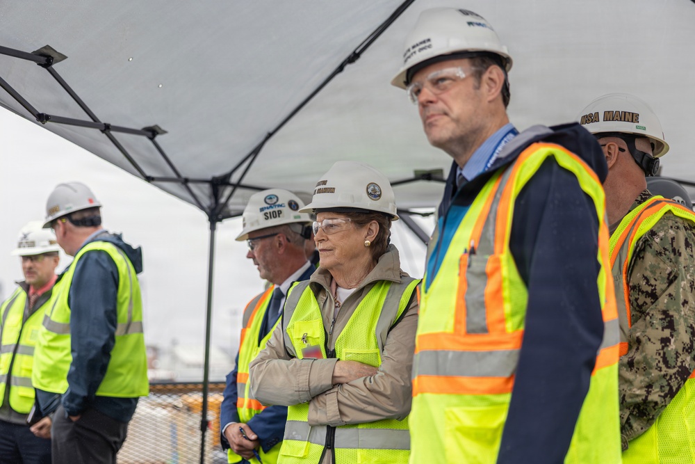A group of individuals wearing white hard hats and high-visibility yellow safety vests are gathered under a canopy tent, likely at a construction or industrial site. The group includes civilians and at least one person in military camouflage uniform. They appear to be observing or attending a formal site visit or inspection event.