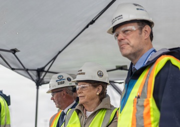 A group of individuals wearing white hard hats and high-visibility yellow safety vests are gathered under a canopy tent, likely at a construction or industrial site. The group includes civilians and at least one person in military camouflage uniform. They appear to be observing or attending a formal site visit or inspection event.