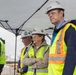 A group of individuals wearing white hard hats and high-visibility yellow safety vests are gathered under a canopy tent, likely at a construction or industrial site. The group includes civilians and at least one person in military camouflage uniform. They appear to be observing or attending a formal site visit or inspection event.